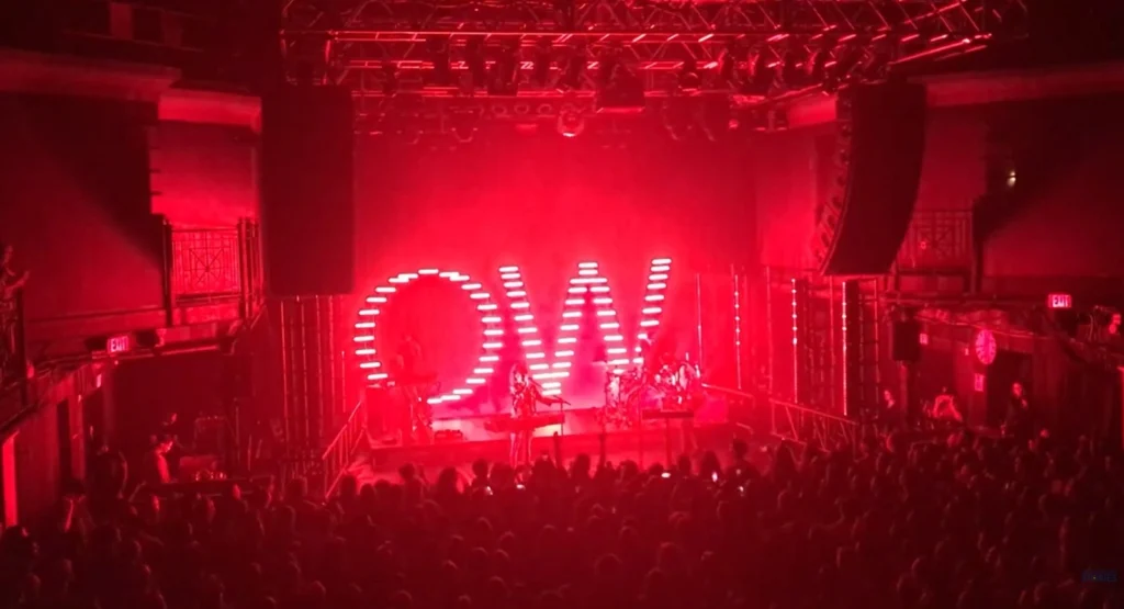 Concert performance under dynamic red lighting with an illuminated 'OW' sign, capturing the energy of the live music experience.